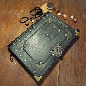 Puede incluir: Un diario encuadernado en cuero negro con detalles en lat&oacute;n y una cerradura. El diario tiene un aspecto desgastado y est&aacute; decorado con dise&ntilde;os ornamentados.