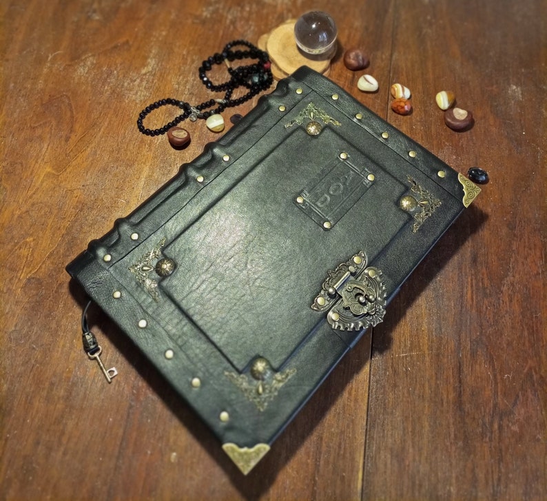 Puede incluir: Un diario encuadernado en cuero negro con detalles en lat&oacute;n y una cerradura. El diario tiene un aspecto desgastado y est&aacute; decorado con dise&ntilde;os ornamentados.