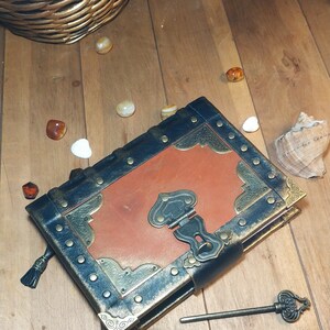May include: A brown leather-bound journal with a black cover and gold accents. The journal has a metal clasp and a keyhole. A small key is lying on the wooden surface next to the journal.
