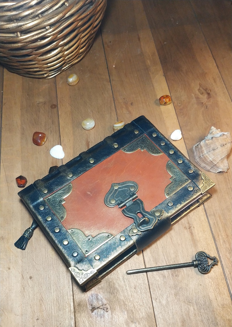 May include: A brown leather-bound journal with a black cover and gold accents. The journal has a metal clasp and a keyhole. A small key is lying on the wooden surface next to the journal.