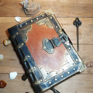 May include: A brown leather-bound journal with a brass lock and key. The journal has a distressed look and is decorated with gold accents. The journal is sitting on a wooden surface.