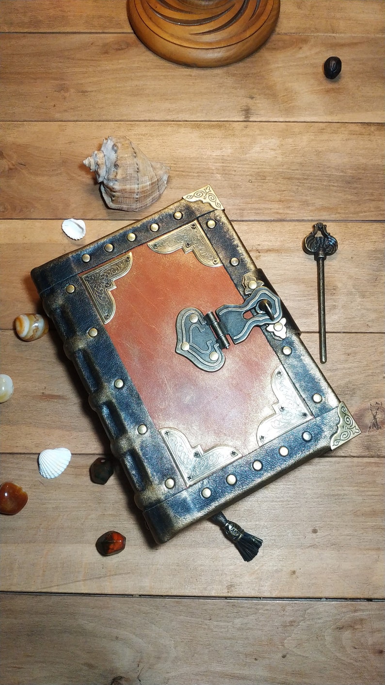 May include: A brown leather-bound journal with a brass lock and key. The journal has a distressed look and is decorated with gold accents. The journal is sitting on a wooden surface.