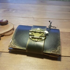 May include: A dark leather journal with a brass clasp and corner accents. The journal is closed with a wide leather strap and a decorative brass clasp. The corners are adorned with ornate brass details. A small tag hangs from the clasp.