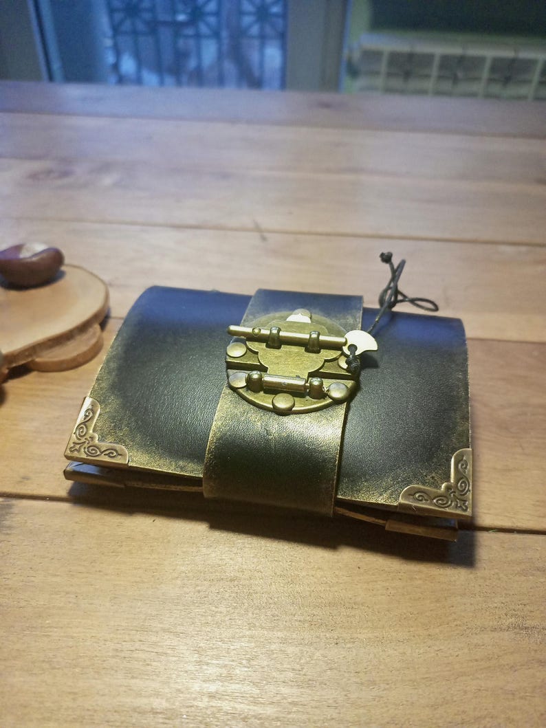May include: A dark leather journal with a brass clasp and corner accents. The journal is closed with a wide leather strap and a decorative brass clasp. The corners are adorned with ornate brass details. A small tag hangs from the clasp.