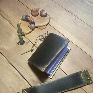 May include: An open black leather journal with gold-colored trim and a matching strap with brass hardware. A beaded bracelet with red and green accents and a small wooden dish with two chestnuts are also present on a wooden surface.
