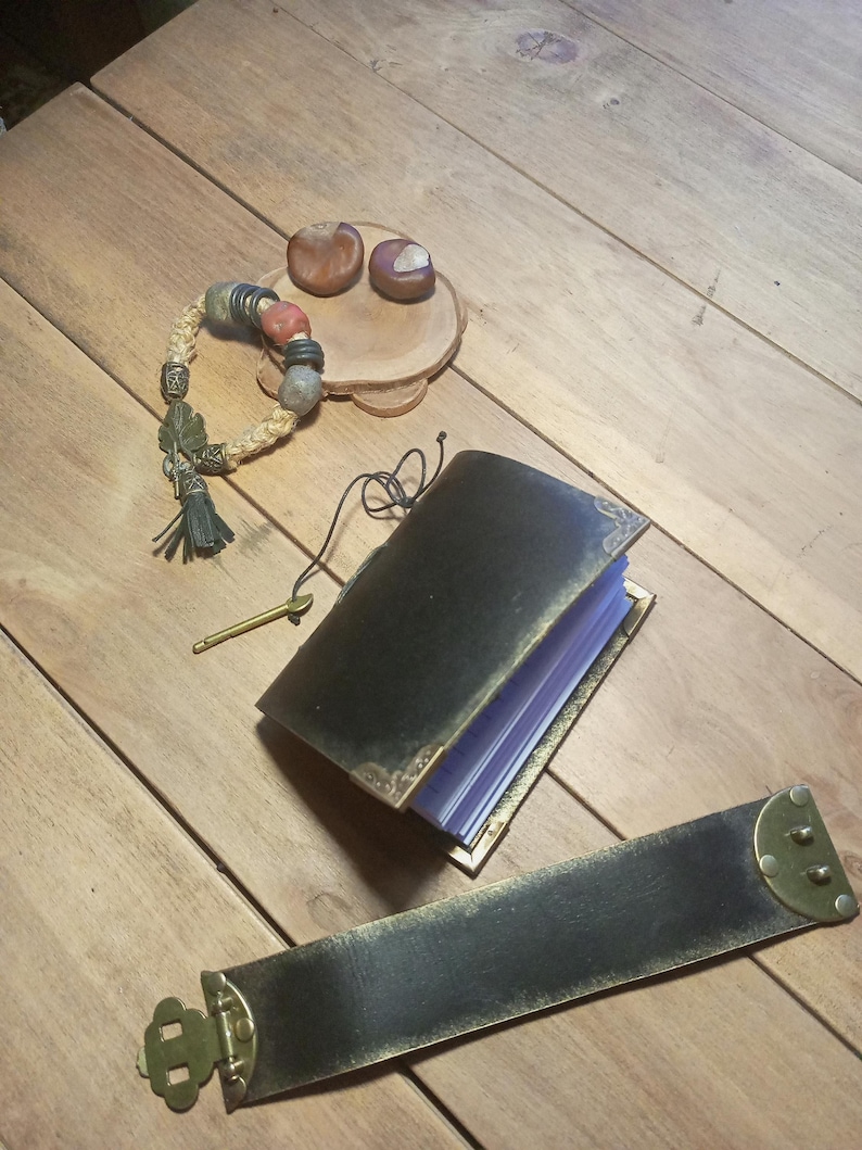 May include: An open black leather journal with gold-colored trim and a matching strap with brass hardware. A beaded bracelet with red and green accents and a small wooden dish with two chestnuts are also present on a wooden surface.