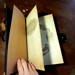 Puede incluir: Un diario encuadernado en cuero negro con un dise&ntilde;o dorado decorativo en la portada. El diario est&aacute; abierto en una p&aacute;gina en blanco con un marco decorativo.