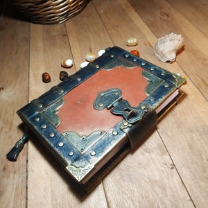 May include: A brown leather journal with a black leather cover and gold metal accents. The journal has a clasp closure and a decorative design on the front cover.