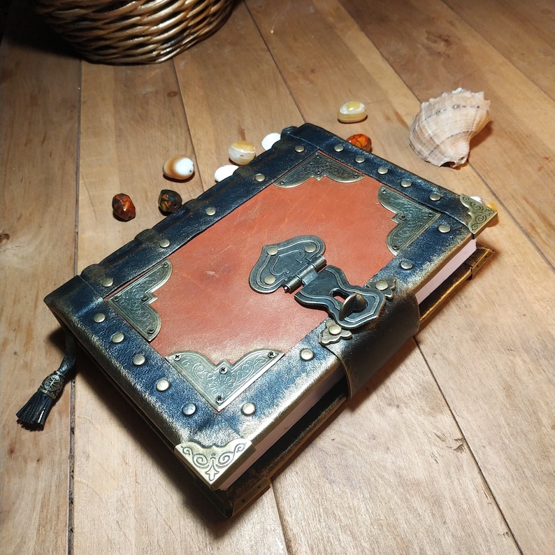 May include: A brown leather journal with a black leather cover and gold metal accents. The journal has a clasp closure and a decorative design on the front cover.