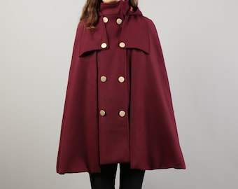Può includere: Un mantello con cappuccio bordeaux con bottoni dorati. Il mantello è doppiopetto e ha un design fluido. La modella indossa leggings neri e stivali marroni.