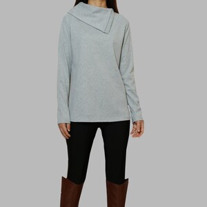 Può includere: Una camicia grigia a maniche lunghe con un collo a scialle e maniche lunghe. La camicia è abbinata a leggings neri e stivali marroni alti al ginocchio.