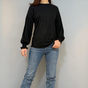 May include: A person wearing a black long-sleeve shirt with ruffled cuffs and blue jeans. The person is also wearing brown boots with buckles.