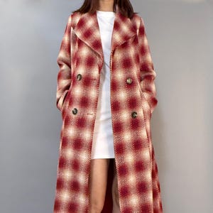 Puede incluir: Un abrigo largo a cuadros rojos y crema con diseño cruzado y botones dorados. El abrigo se lleva sobre un vestido blanco y se combina con tacones negros. La longitud del abrigo llega hasta los tobillos.