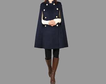 Pode incluir: Uma capa azul marinho com botões dourados e gola alta. A capa é usada sobre uma camisa branca de manga comprida e leggings pretos. A pessoa está usando botas marrons.