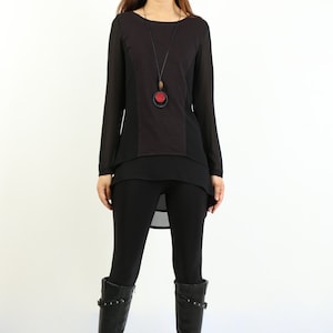 Könnte beinhalten: Ein schwarzes Langarm-Tunika-Oberteil mit einem geschichteten Design und einer schwarzen Anhänger-Halskette mit einem roten Kreis. Das Oberteil wird mit schwarzen Leggings und schwarzen Stiefeln kombiniert.
