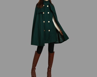 Può includere: Un mantello doppiopetto verde scuro con colletto alto e sei grandi bottoni color oro. Il capo è indossato sopra una maglia a maniche lunghe color crema, leggings neri e stivali alti in pelle marrone fino al ginocchio.