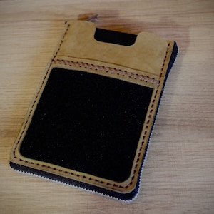 May include: A tan leather zippered pouch with a black fabric interior. The pouch has a stitched design around the edges.