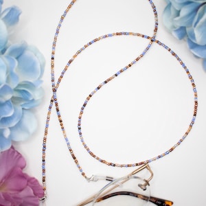 May include: A beaded eyeglass chain with small, colorful beads in shades of blue, brown, and gold. The chain is draped over a pair of eyeglasses with a silver clasp. The background includes blue and purple flowers.