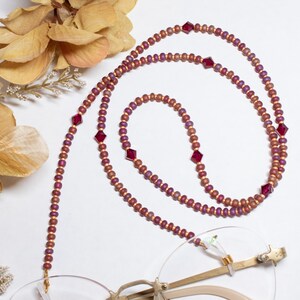 May include: A beaded eyeglass chain with a mix of small, round, and larger, square-cut beads. The chain features a color palette of burgundy, gold, and red, and is draped over a pair of gold-rimmed glasses. The background is white.