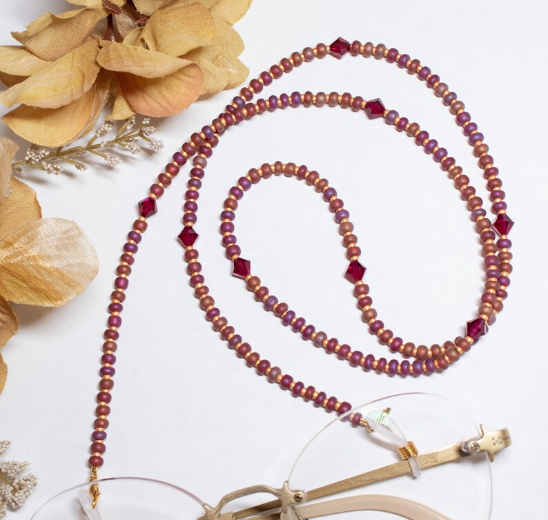May include: A beaded eyeglass chain with a mix of small, round, and larger, square-cut beads. The chain features a color palette of burgundy, gold, and red, and is draped over a pair of gold-rimmed glasses. The background is white.