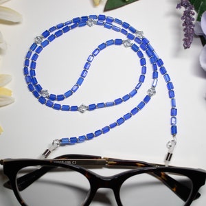 May include: A blue beaded eyeglass chain with rectangular beads and silver accents. The chain is draped over a pair of tortoiseshell glasses. Decorative flowers in yellow and purple are in the background.