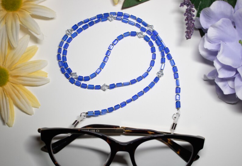 May include: A blue beaded eyeglass chain with rectangular beads and silver accents. The chain is draped over a pair of tortoiseshell glasses. Decorative flowers in yellow and purple are in the background.