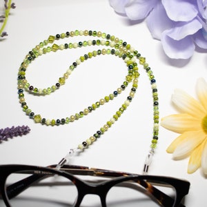 May include: A beaded eyeglass chain with a mix of green, yellow, and cream-colored beads. The chain is draped over a pair of tortoiseshell glasses. The background includes flowers in shades of purple and yellow.