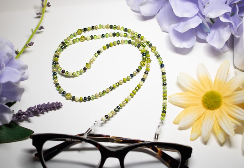 May include: A beaded eyeglass chain with a mix of green, yellow, and cream-colored beads. The chain is draped over a pair of tortoiseshell glasses. The background includes flowers in shades of purple and yellow.