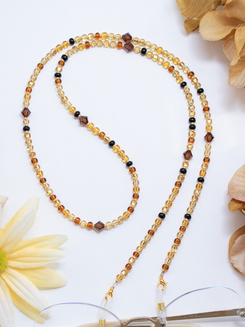 May include: A beaded eyeglass chain in shades of gold, amber, and black. The chain features small, round beads interspersed with faceted, square beads. The chain is attached to a pair of eyeglasses with gold-colored hardware.