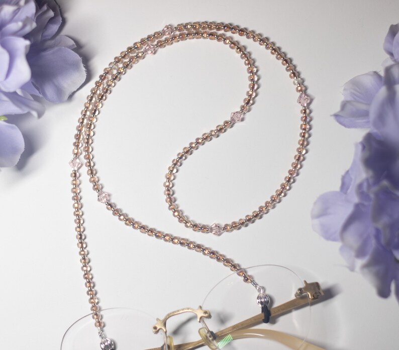 May include: A beaded eyeglass chain with small, round, copper-colored beads and interspersed pink crystal beads. The chain is draped over a pair of eyeglasses with gold frames. The background features soft purple flowers.