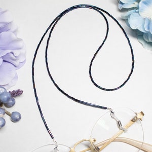 May include: A beaded eyeglass chain with iridescent dark blue and green beads. The chain is attached to a pair of eyeglasses with a gold frame. The background is white with purple and blue floral accents.