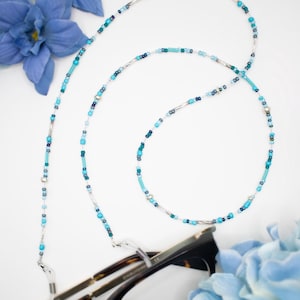 May include: A beaded eyeglass chain in shades of turquoise, teal, and silver. The chain is draped over a pair of tortoiseshell eyeglasses. Blue flowers are in the background.