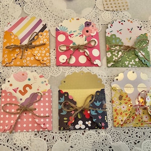 May include: A collection of six small, decorative paper envelopes with scalloped edges, tied with twine. The envelopes feature various patterns, including polka dots, floral designs, and stripes. Pink paper roses are scattered around the envelopes.