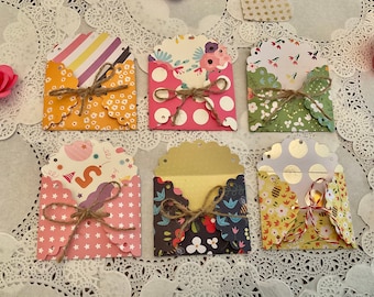 May include: A collection of six small, decorative paper envelopes with scalloped edges, tied with twine. The envelopes feature various patterns, including polka dots, floral designs, and stripes. Pink paper roses are scattered around the envelopes.