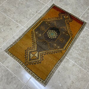 Könnte beinhalten: Ein kleiner, rechteckiger Teppich mit braunem und beigefarbenem Hintergrund und einem dunkelbraunen geometrischen Muster in der Mitte. Der Teppich hat einen weißen Fransenrand.