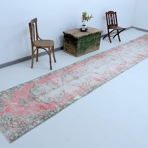 Puede incluir: Una alfombra larga y rectangular con un patrón floral rosa y verde. La alfombra está colocada sobre un suelo blanco con dos sillas de madera y un cofre de madera en el fondo.