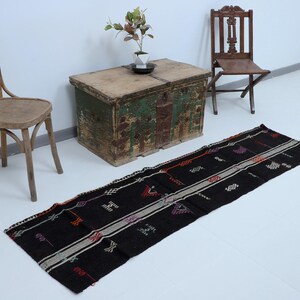 May include: A black runner rug with geometric patterns and colourful accents. The rug is displayed on a white floor, with two wooden chairs and a rustic wooden chest in the background.