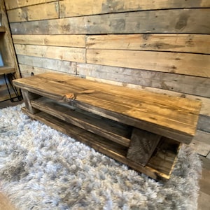 Op de afbeelding: Een rustieke houten bank met twee planken. De bank is gemaakt van gerecycled hout met een natuurlijke afwerking die de houtnerf benadrukt. De bank staat op een zacht, grijs tapijt. De achtergrond is een houten wand. Ongeveer 120 cm lang.