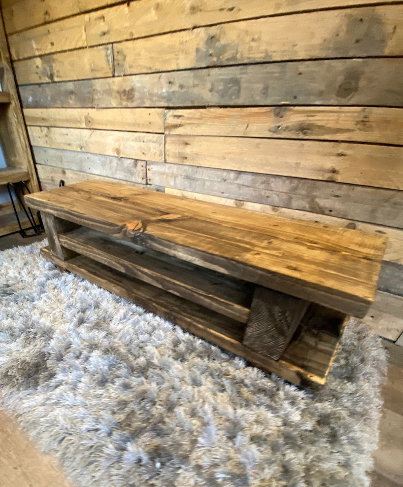 Op de afbeelding: Een rustieke houten bank met twee planken. De bank is gemaakt van gerecycled hout met een natuurlijke afwerking die de houtnerf benadrukt. De bank staat op een zacht, grijs tapijt. De achtergrond is een houten wand. Ongeveer 120 cm lang.