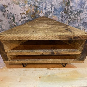 K&ouml;nnte beinhalten: Eckregal aus Altholz in dreieckiger Form. Es hat zwei offene Ablagefl&auml;chen und steht auf schwarzen Metall-Haarnadelbeinen. Das Holz hat eine nat&uuml;rliche, verwitterte Optik mit sichtbarer Maserung und Astl&ouml;chern.