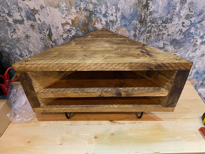 K&ouml;nnte beinhalten: Eckregal aus Altholz in dreieckiger Form. Es hat zwei offene Ablagefl&auml;chen und steht auf schwarzen Metall-Haarnadelbeinen. Das Holz hat eine nat&uuml;rliche, verwitterte Optik mit sichtbarer Maserung und Astl&ouml;chern.