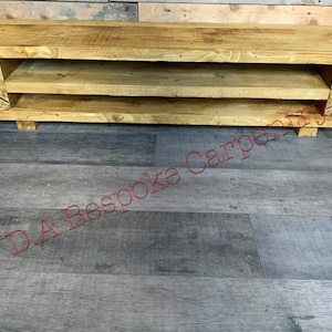 Op de afbeelding: Een rustieke tv-meubel van lichtkleurig hout met twee open planken en een bovenblad. De console heeft een eenvoudig, rechthoekig ontwerp. Geschikt voor een woonkamer of entertainmentruimte. Meet ongeveer 120 cm lang.