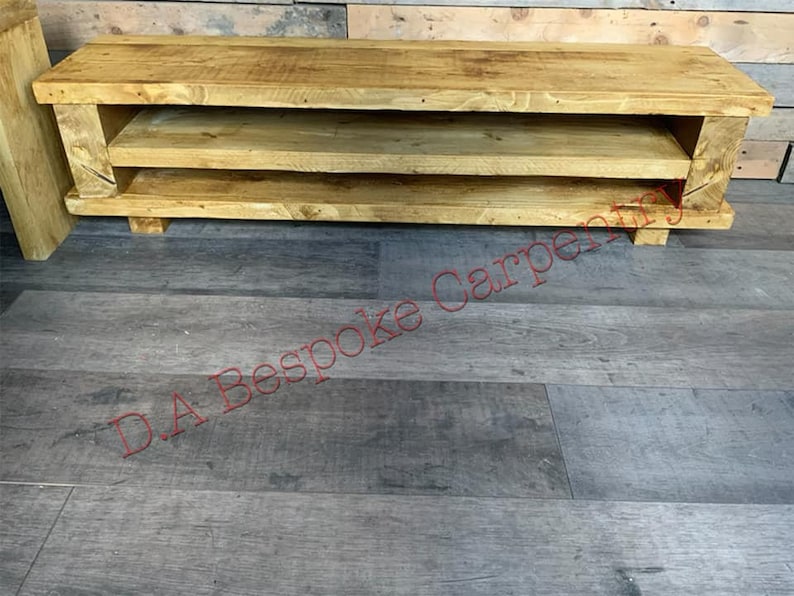 Op de afbeelding: Een rustieke tv-meubel van lichtkleurig hout met twee open planken en een bovenblad. De console heeft een eenvoudig, rechthoekig ontwerp. Geschikt voor een woonkamer of entertainmentruimte. Meet ongeveer 120 cm lang.