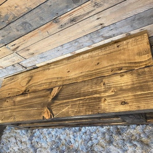 Op de afbeelding: Een rustieke, rechthoekige salontafel van gerecycled hout. De tafel heeft een donkerbruine beits, die de natuurlijke houtnerf en knopen laat zien. De tafel wordt ondersteund door zwarte metalen haarspeldpoten. De tafel is ongeveer 122 cm lang.