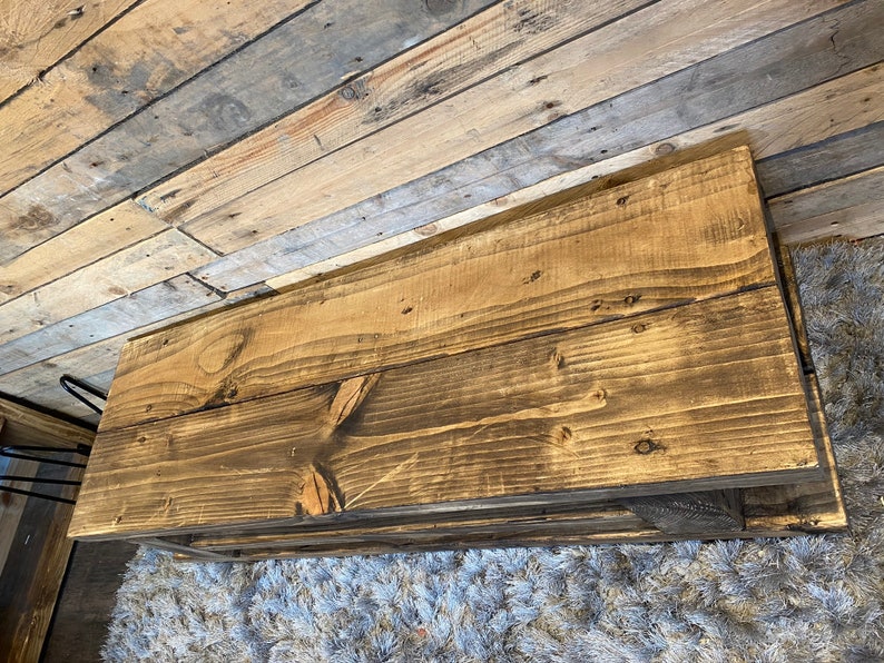 Op de afbeelding: Een rustieke, rechthoekige salontafel van gerecycled hout. De tafel heeft een donkerbruine beits, die de natuurlijke houtnerf en knopen laat zien. De tafel wordt ondersteund door zwarte metalen haarspeldpoten. De tafel is ongeveer 122 cm lang.