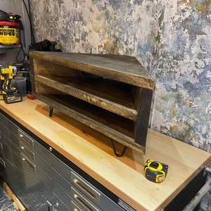 K&ouml;nnte beinhalten: Rustikales, eckf&ouml;rmiges Holzregal mit drei Ebenen. Das Regal ist aus dunkel gebeiztem Holz gefertigt und hat schwarze Haarnadelbeine. Ein gelb-schwarzes Ma&szlig;band liegt daneben. Das Regal ist f&uuml;r eine Ecke konzipiert.