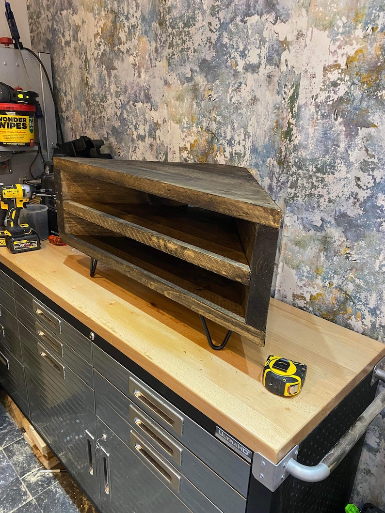 K&ouml;nnte beinhalten: Rustikales, eckf&ouml;rmiges Holzregal mit drei Ebenen. Das Regal ist aus dunkel gebeiztem Holz gefertigt und hat schwarze Haarnadelbeine. Ein gelb-schwarzes Ma&szlig;band liegt daneben. Das Regal ist f&uuml;r eine Ecke konzipiert.