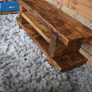 Op de afbeelding: Een rustieke houten bank met een rijke bruine afwerking. De bank heeft twee planken en is gemaakt van dikke houten planken. Hij staat op een grijze, harige vloerkleed. De bank is ongeveer 122 cm lang.
