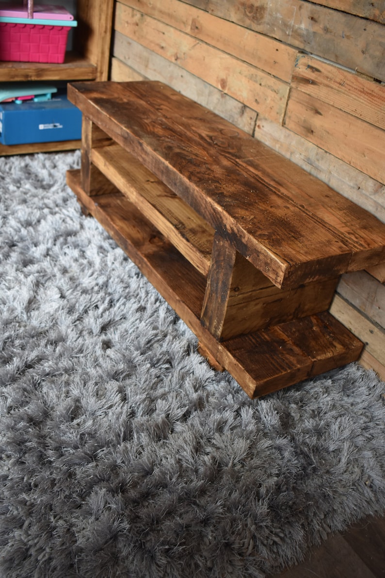 Op de afbeelding: Een rustieke houten bank met een rijke bruine afwerking. De bank heeft twee planken en is gemaakt van dikke houten planken. Hij staat op een grijze, harige vloerkleed. De bank is ongeveer 122 cm lang.