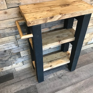 May include: A rustic wooden side table with a natural wood top and two shelves. The legs are painted black, and a wooden handle is attached to the side. The table is made from reclaimed wood and has a distressed finish.